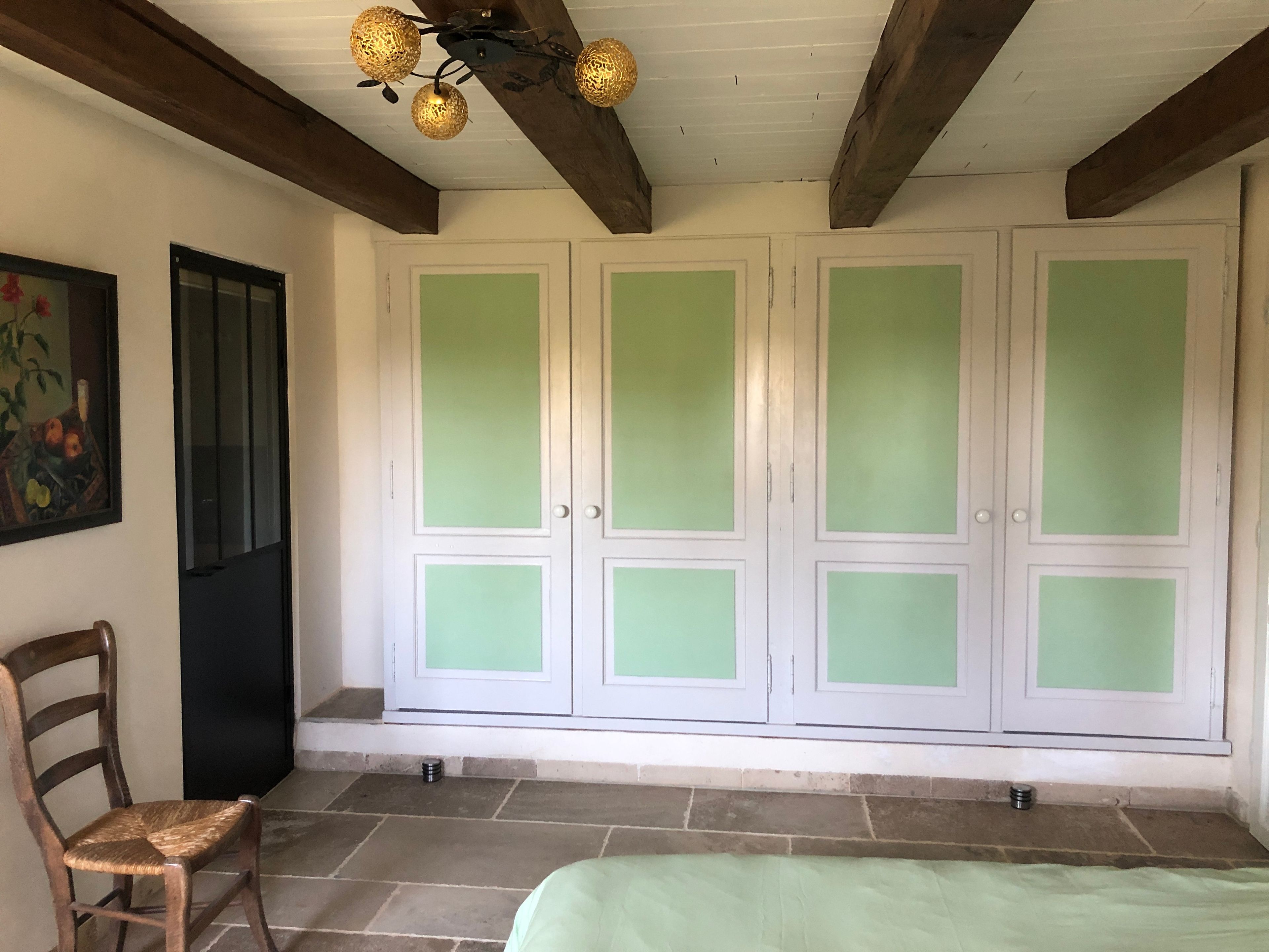A room with a bed, a chair, a wardrobe with frosted glass doors, a ceiling light fixture, and a painting on the wall.