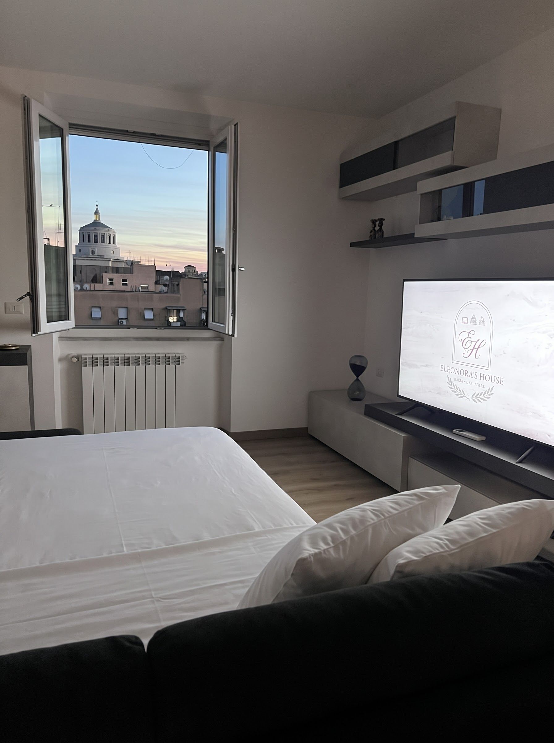 Une chambre à coucher avec un lit, une fenêtre donnant sur des immeubles, un radiateur et un téléviseur posé sur un meuble.