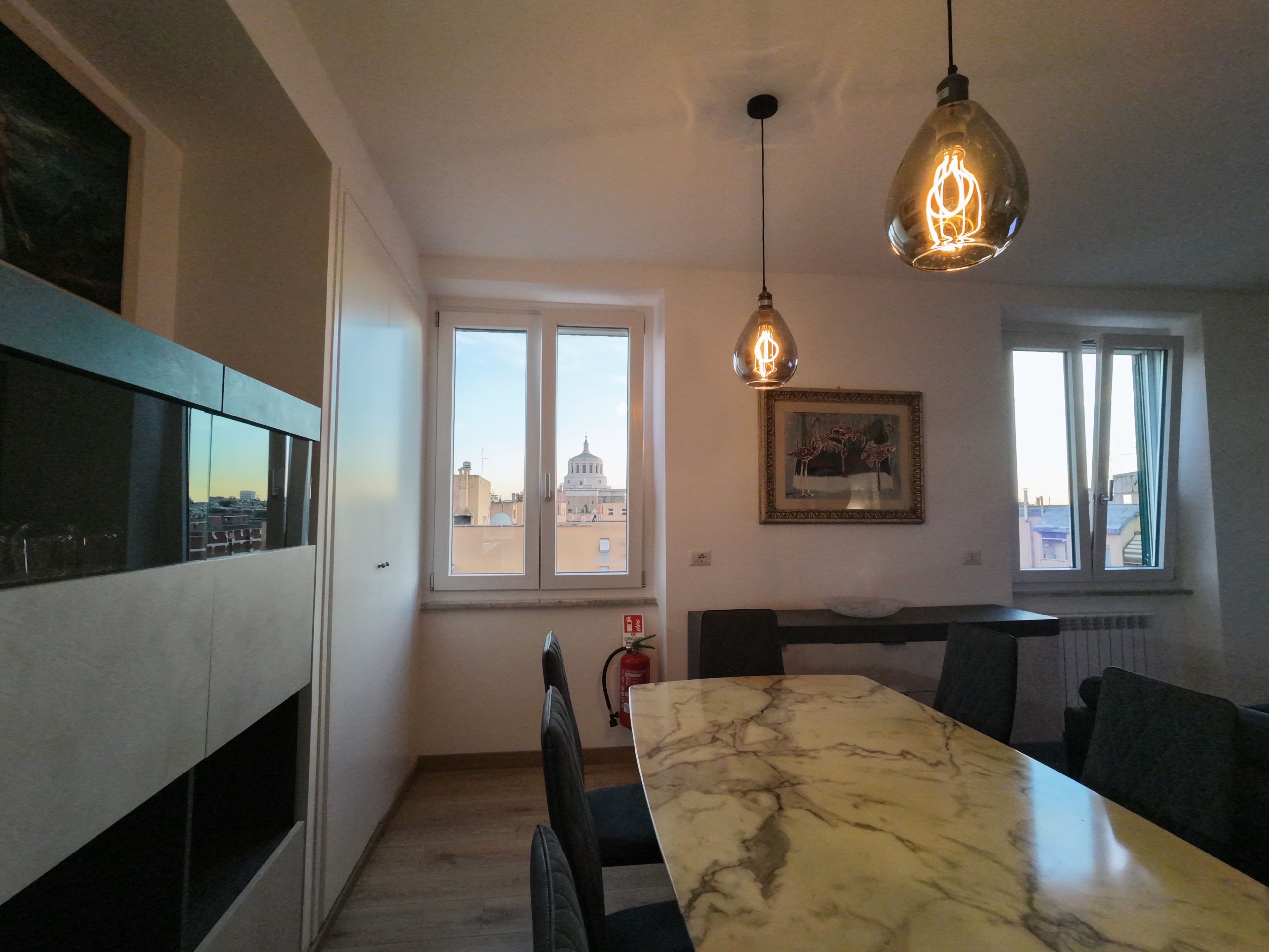 Un coin repas avec une table en marbre, des chaises noires, un luminaire suspendu et une fenêtre donnant sur la ville.