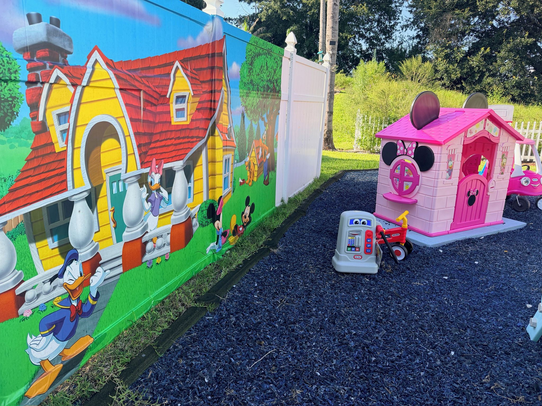 Ein farbenfrohes Wandgemälde mit Cartoon-Figuren, ein kleines rosa Spielhäuschen und ein Spielzeugauto auf einem Schotterweg.