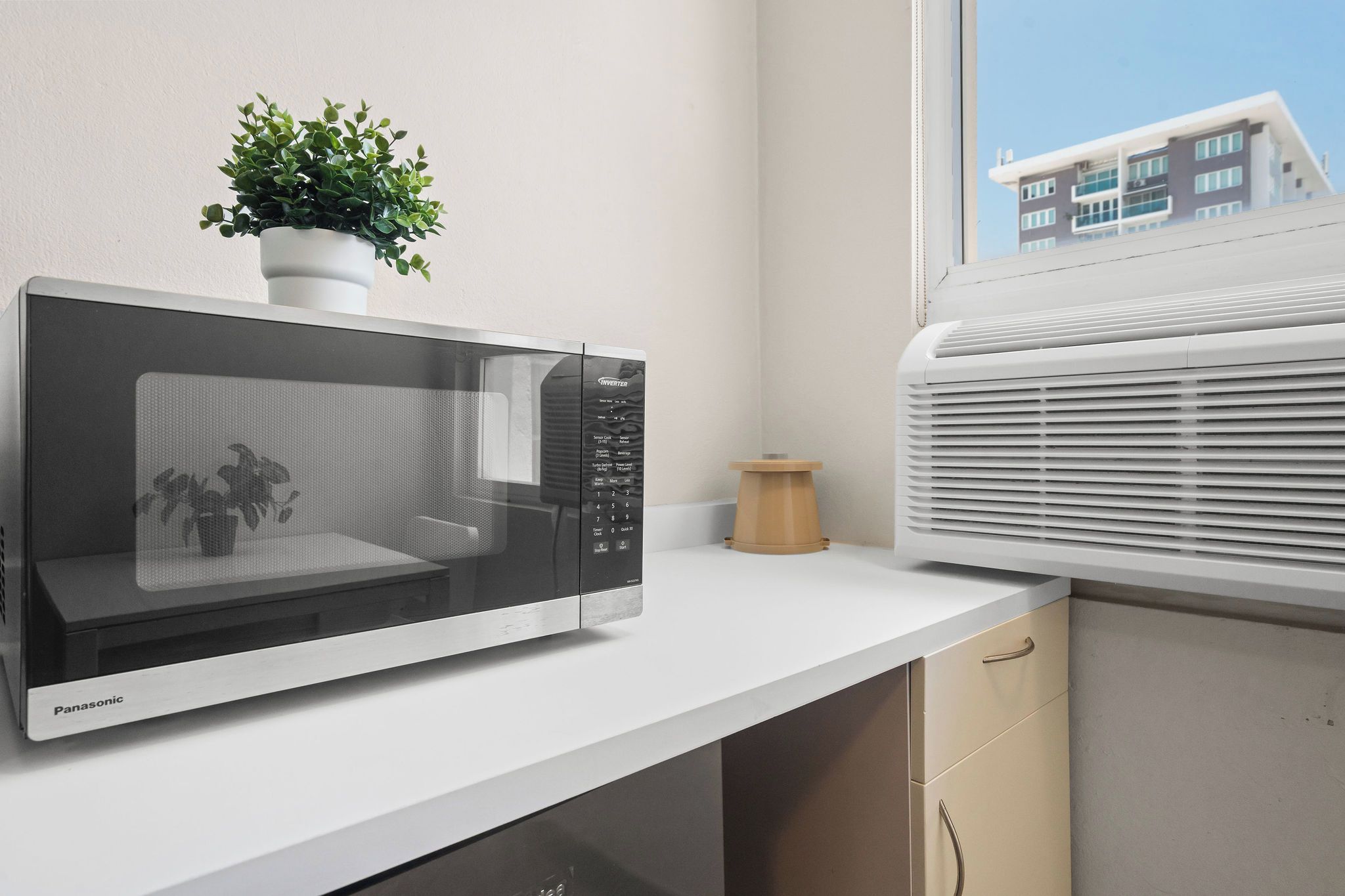 A Panasonic microwave oven on a white countertop with a potted plant, a wooden stool, and a window with a view of a building.