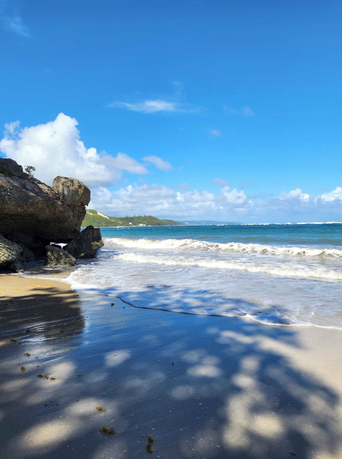 A beach with clear skies, waves, and a rocky outcrop.