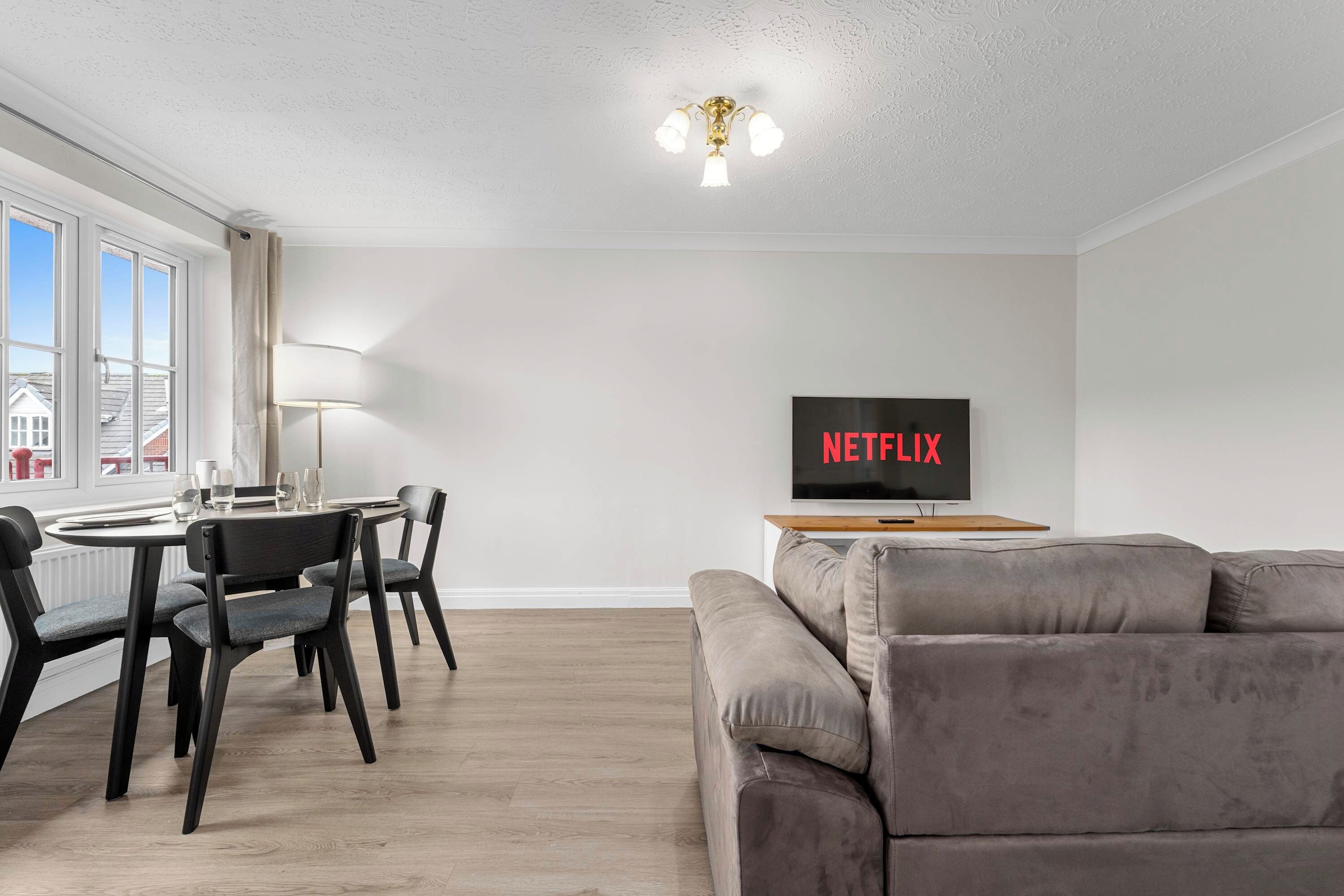 Een moderne woonkamer met een grijze bank, een eettafel met stoelen en een tv waarop 'NETFLIX' staat.