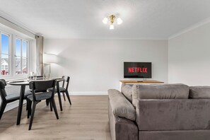 Ein modernes Wohnzimmer mit einem grauen Sofa, einem Esstisch mit Stühlen und einem Fernseher, der 'NETFLIX' anzeigt.