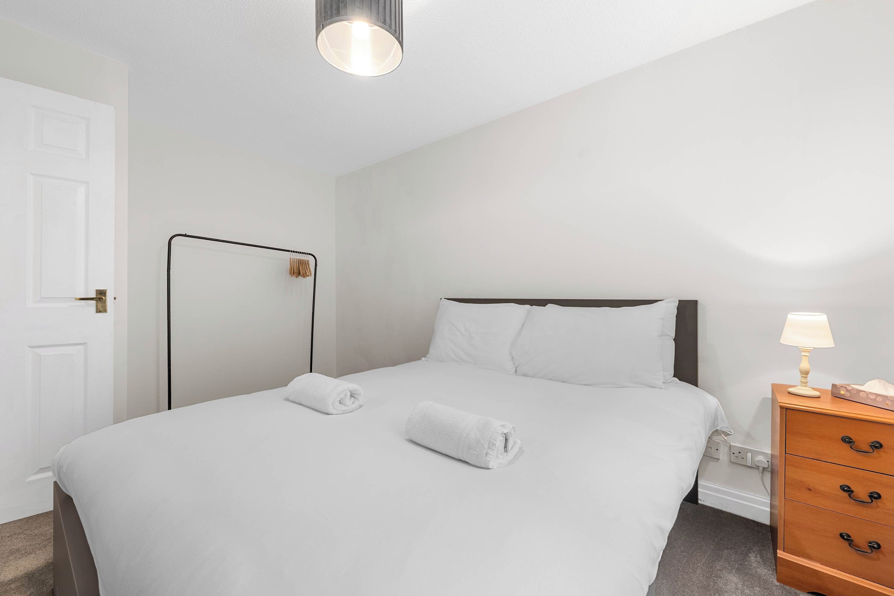 A neatly made bed with white linens and two rolled towels, a bedside table with a lamp, and a wooden dresser with drawers.