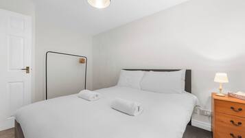 A neatly made bed with white linens and two rolled towels, a bedside table with a lamp, and a wooden dresser with drawers.