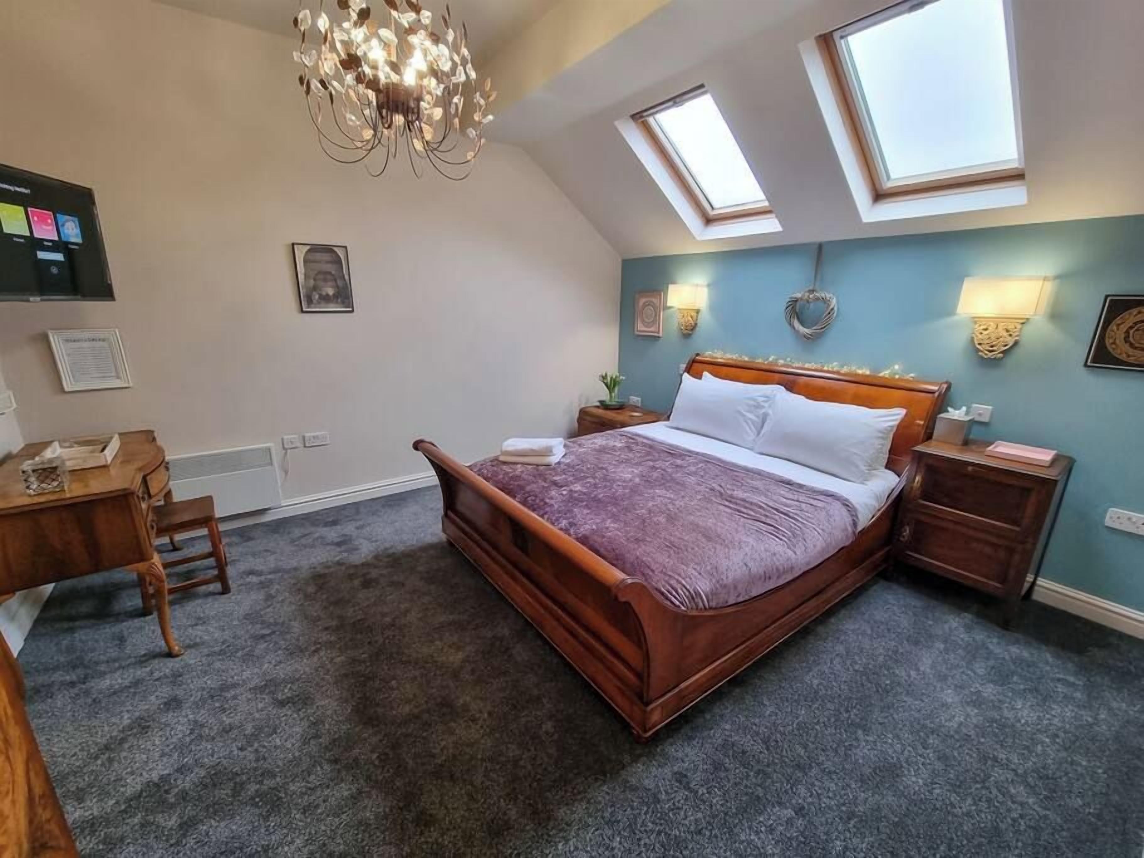 Quarto com cama de madeira, escrivaninha e cadeira. Há duas luminárias na parede e um lustre no teto.