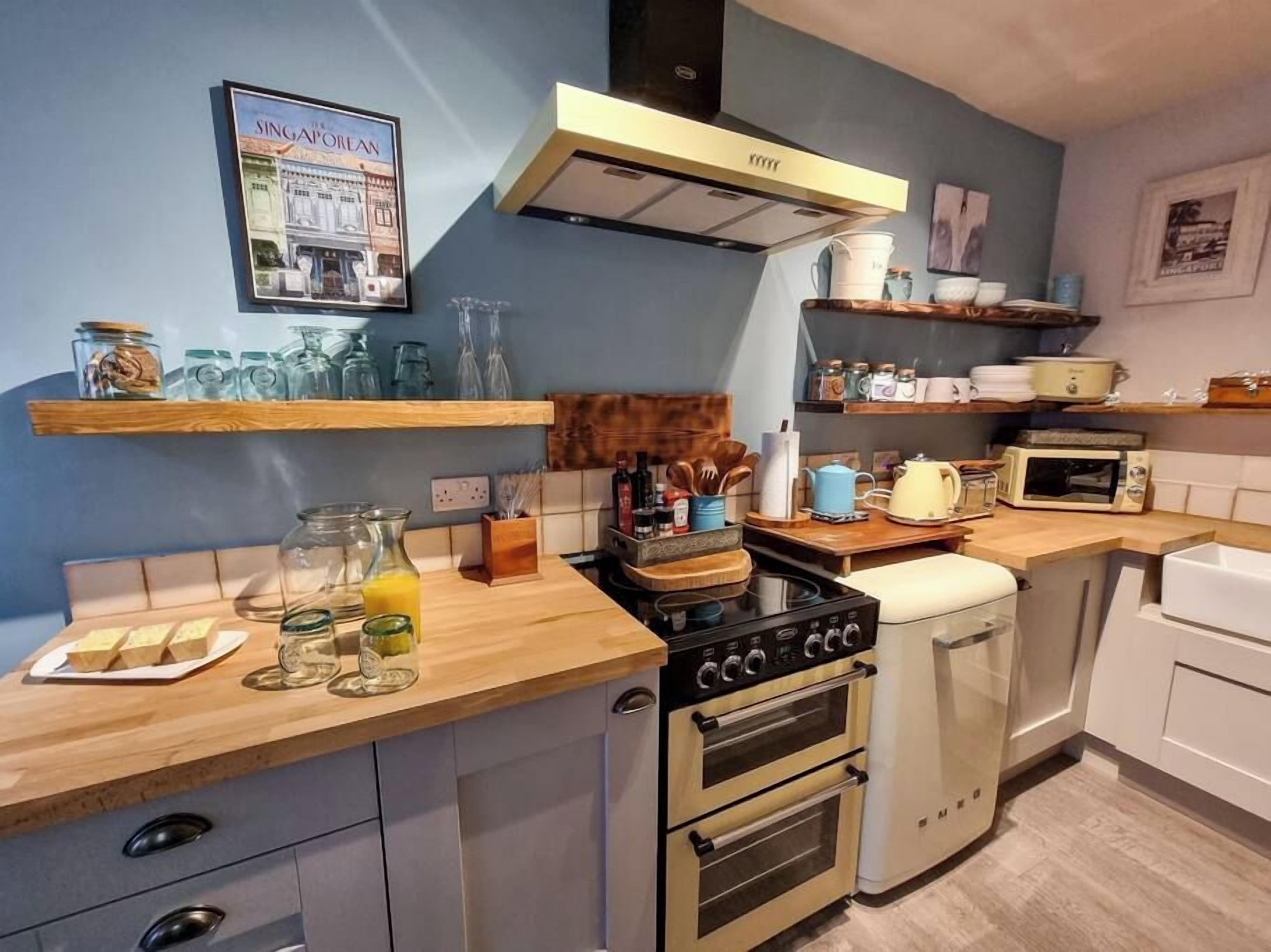 Een compacte keuken met een gasfornuis, oven en magnetron. Er zijn houten planken met potten en keukengerei, en een ingelijst foto aan de muur.