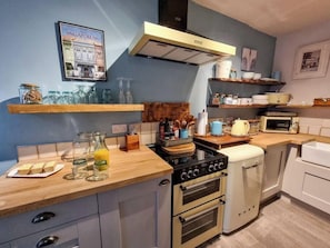 Une cuisine compacte équipée d’une cuisinière à gaz, d’un four et d’un four à micro-ondes. On y trouve des étagères en bois où sont rangés des bocaux et des ustensiles de cuisine, ainsi qu’un tableau encadré accroché au mur.