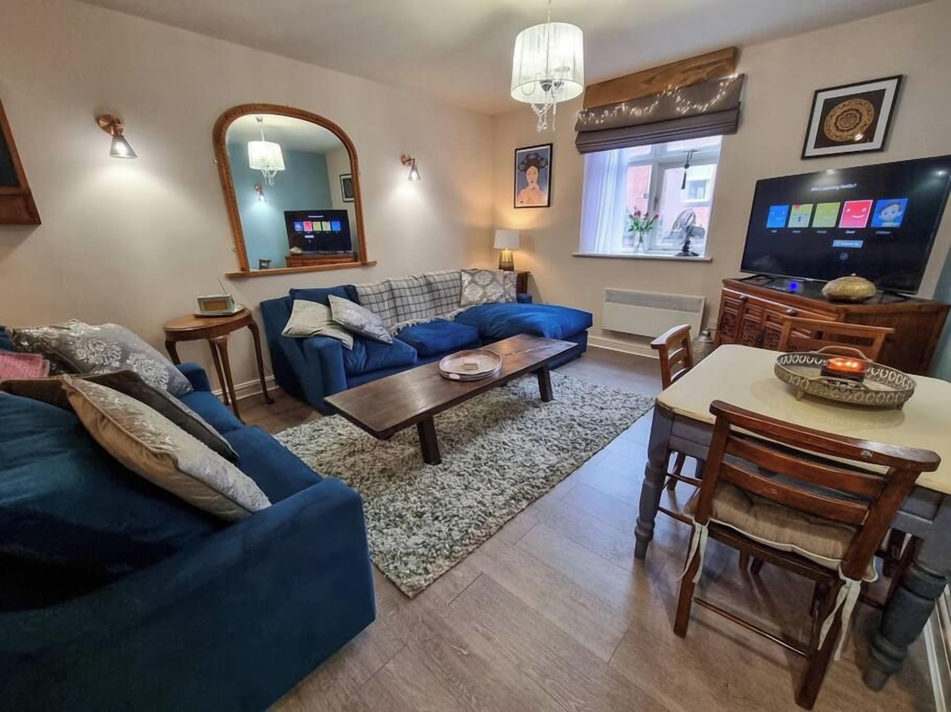 A living room with a blue sofa, a wooden coffee table, a dining table with chairs, a TV, and a large mirror.