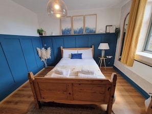 A bedroom with a wooden bed, a blue wall, a window with yellow curtains, and a hanging light fixture. - Flat Next to Piccadilly Station | City Centre (Manchester)