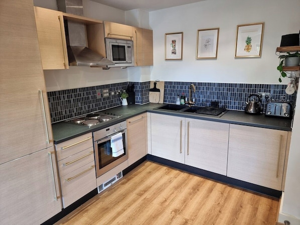 A modern kitchen with a microwave, stove, oven, and sink. There are framed pictures on the wall and a plant on the countertop. - Flat Next to Piccadilly Station | City Centre (Manchester)