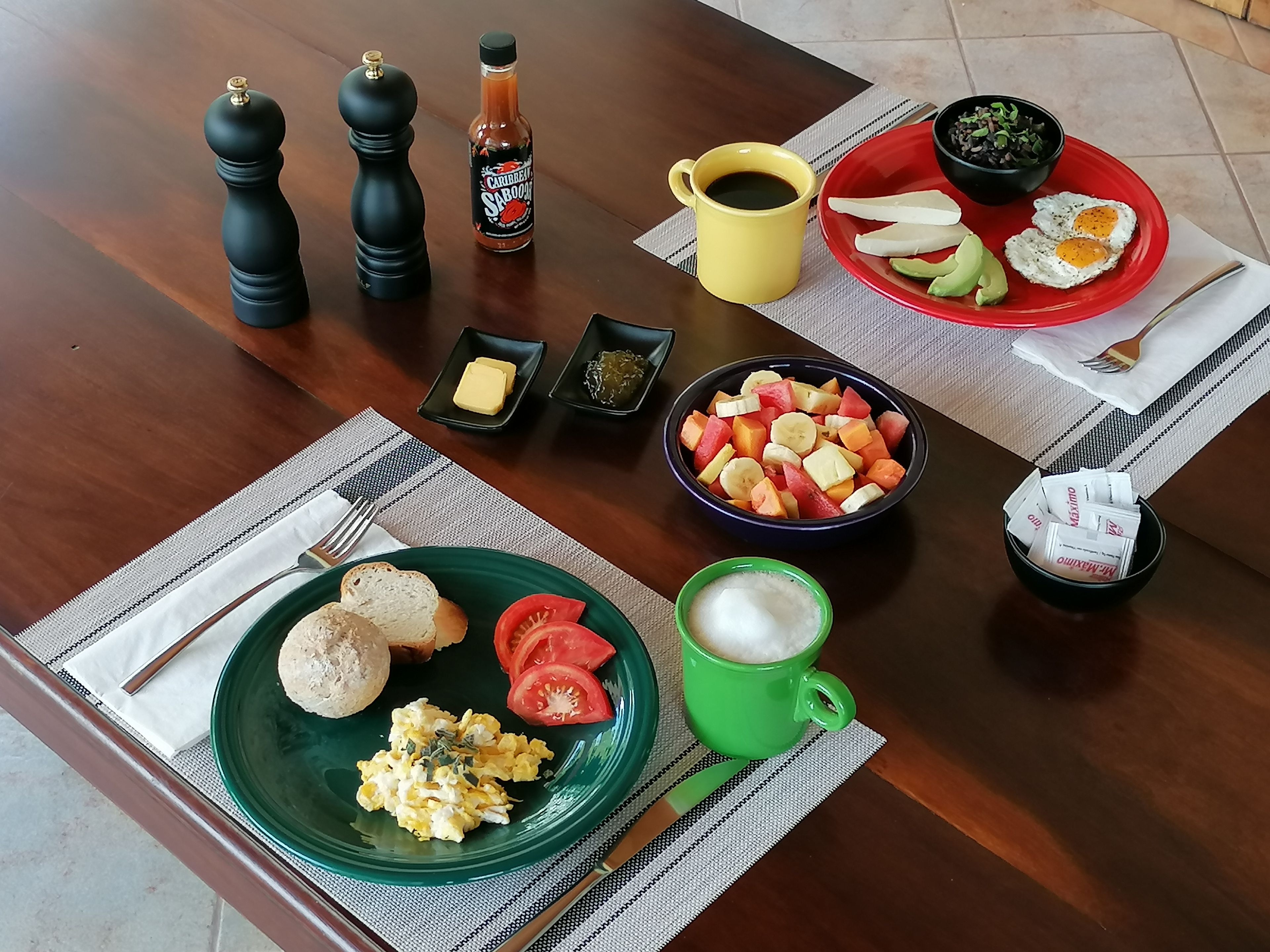 Una mesa de desayuno con un plato de huevos revueltos, tostadas y tomates, un bol de fruta, una taza de café verde, un plato rojo con huevos y verduras, una taza amarilla y condimentos.