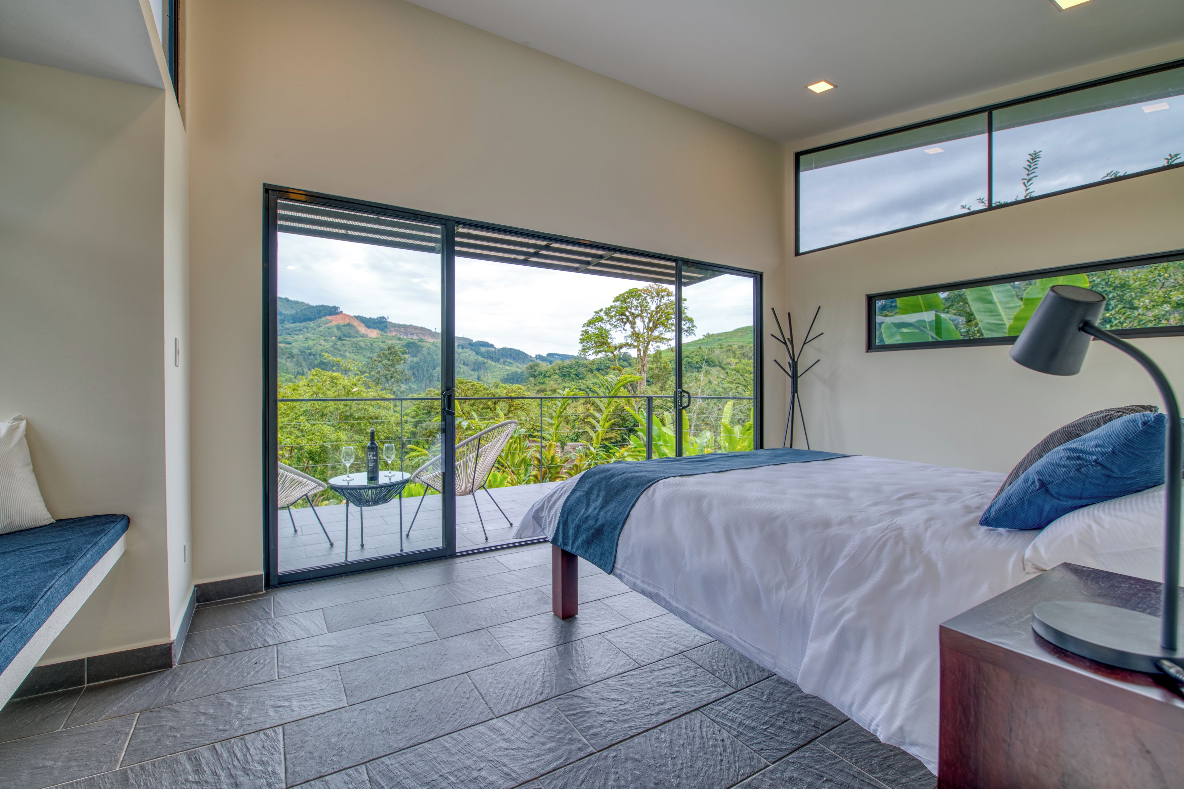 A bedroom with a large sliding glass door leading to a balcony, a bed with a blue accent pillow, a bedside table with a lamp, and a view of a lush landscape.