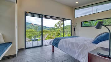 A bedroom with a large sliding glass door leading to a balcony, a bed with a blue accent pillow, a bedside table with a lamp, and a view of a lush landscape.