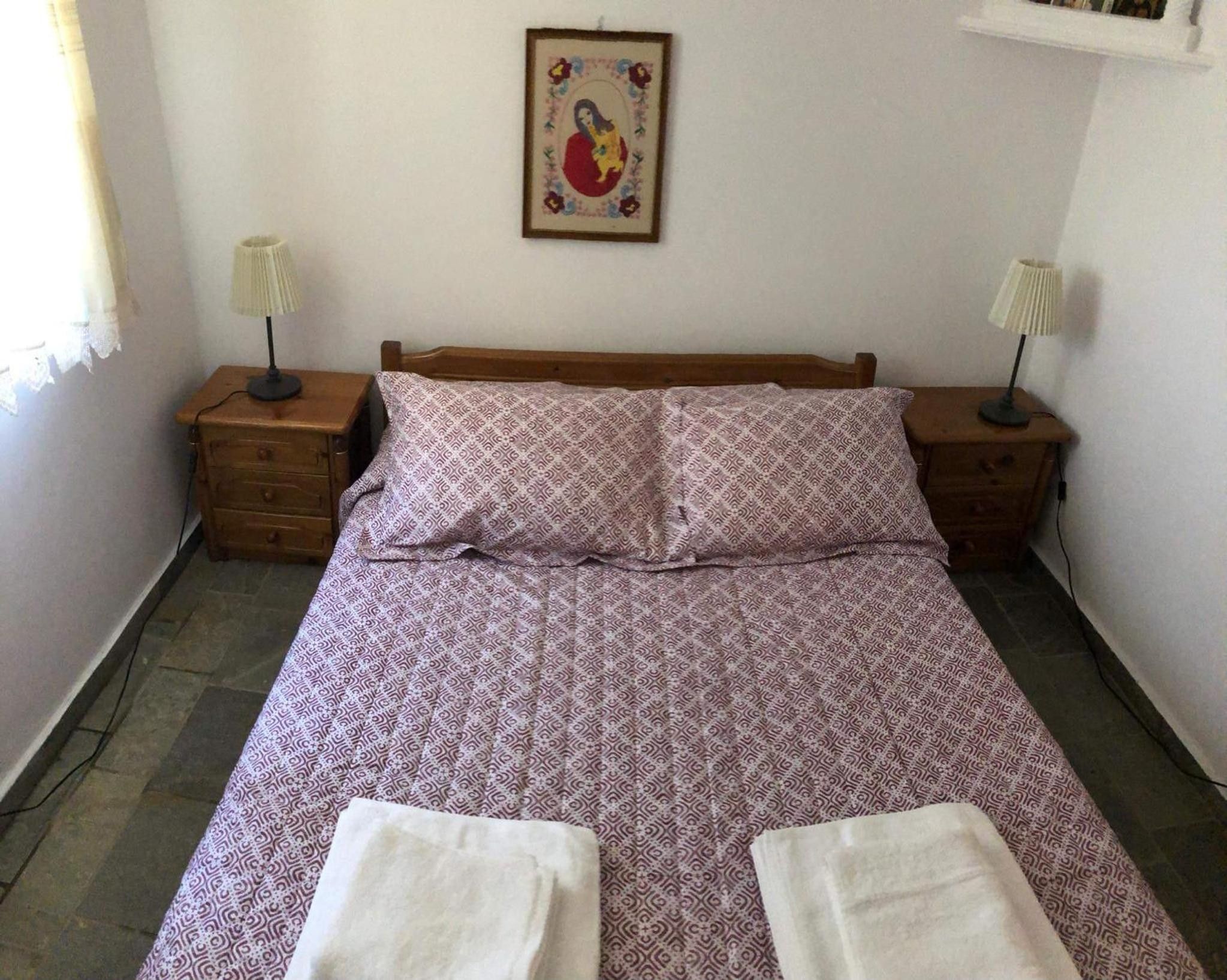 Ein Schlafzimmer mit einem Bett, zwei Nachttischen, einem Wandbild und einem Fenster mit Vorhängen.