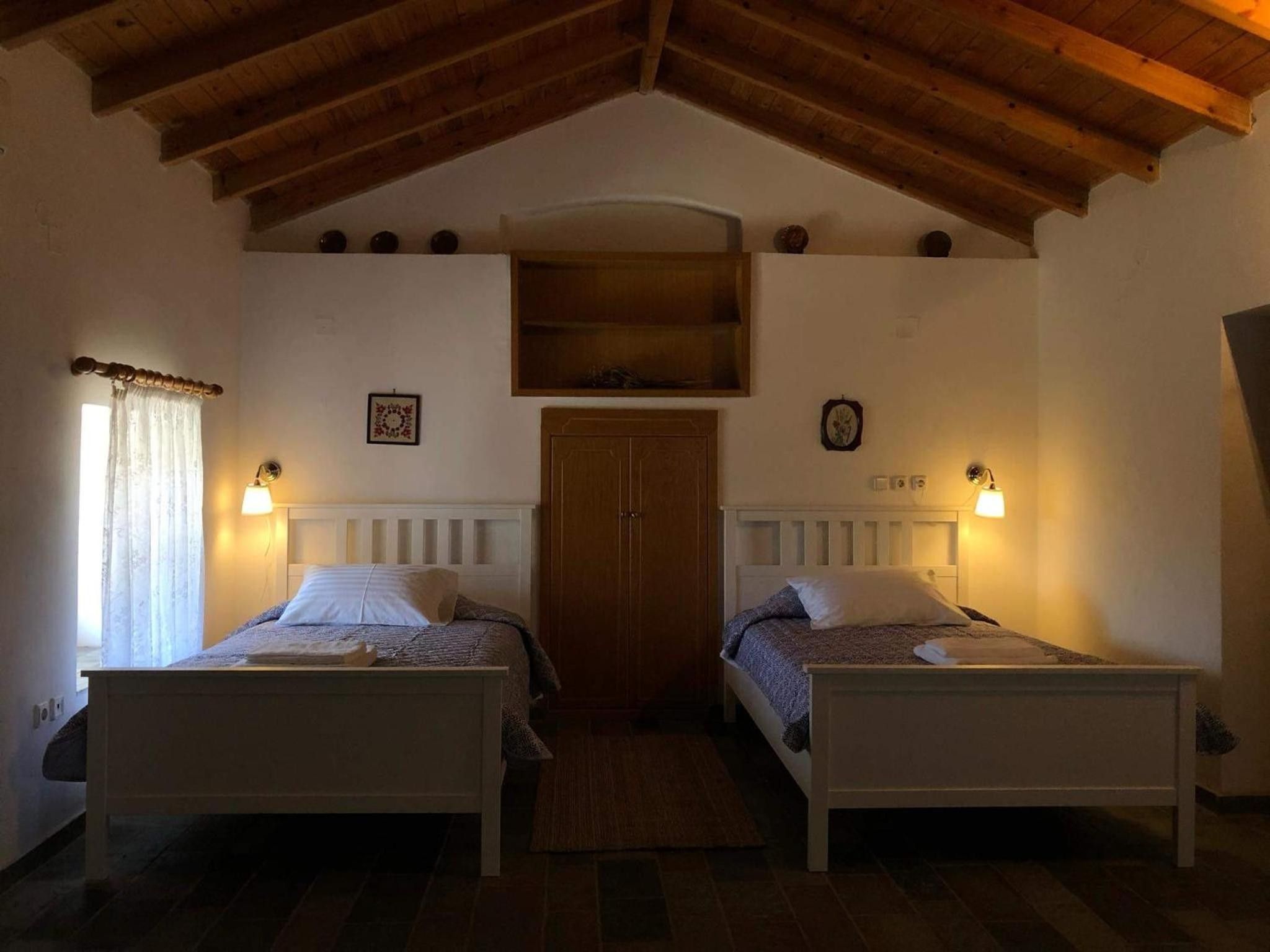 A bedroom with two single beds, a wooden ceiling, a small window with curtains, and wall-mounted lights.