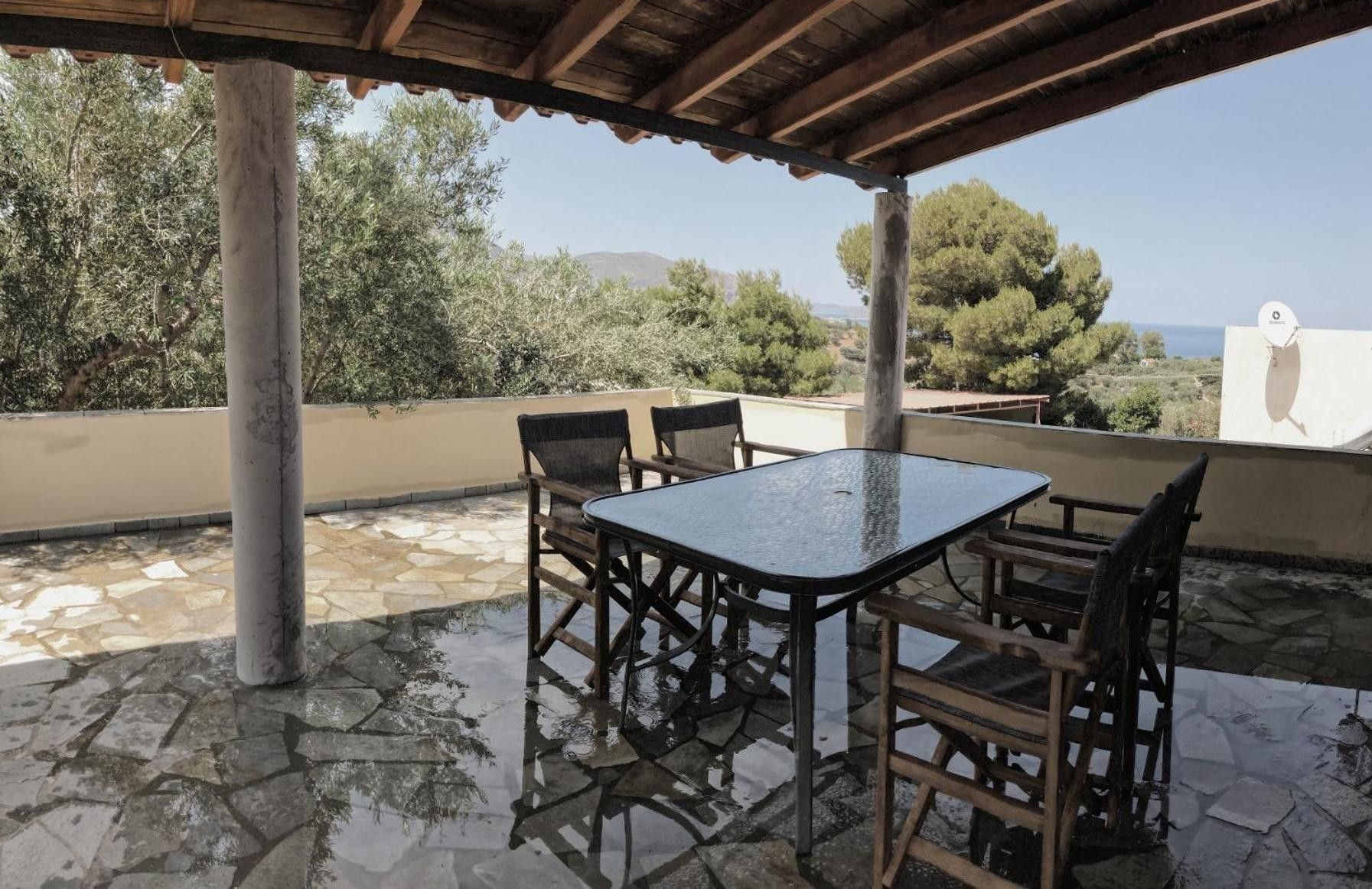 An outdoor patio with a table and chairs, stone flooring, and a view of trees and the sea.