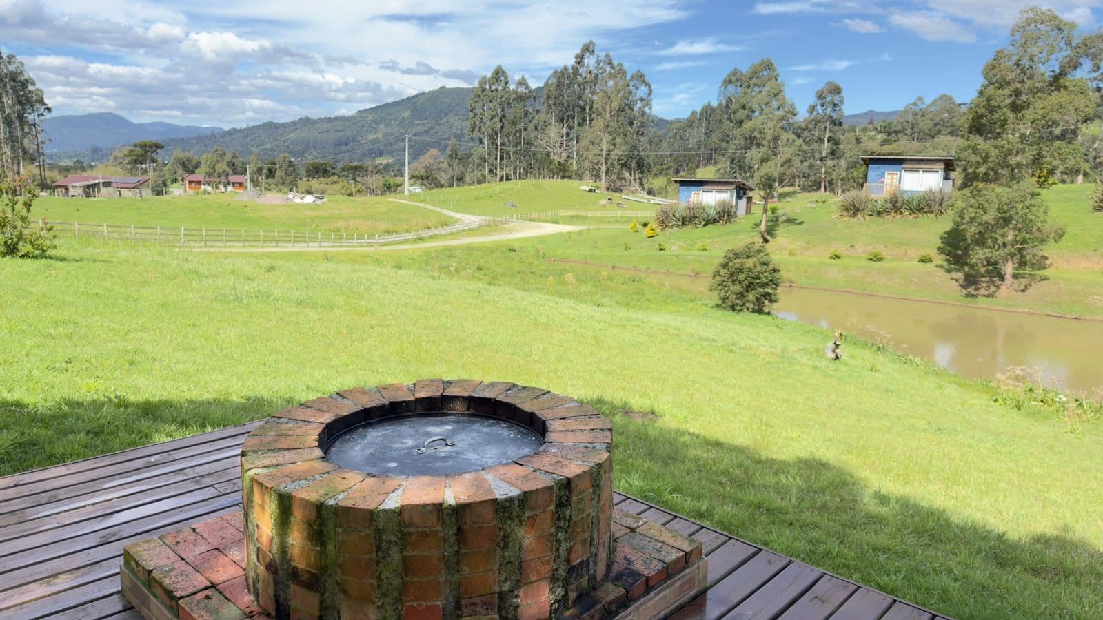 Un pozo de fuego circular al aire libre sobre una terraza de madera, con un campo cubierto de césped y un pequeño estanque al fondo.