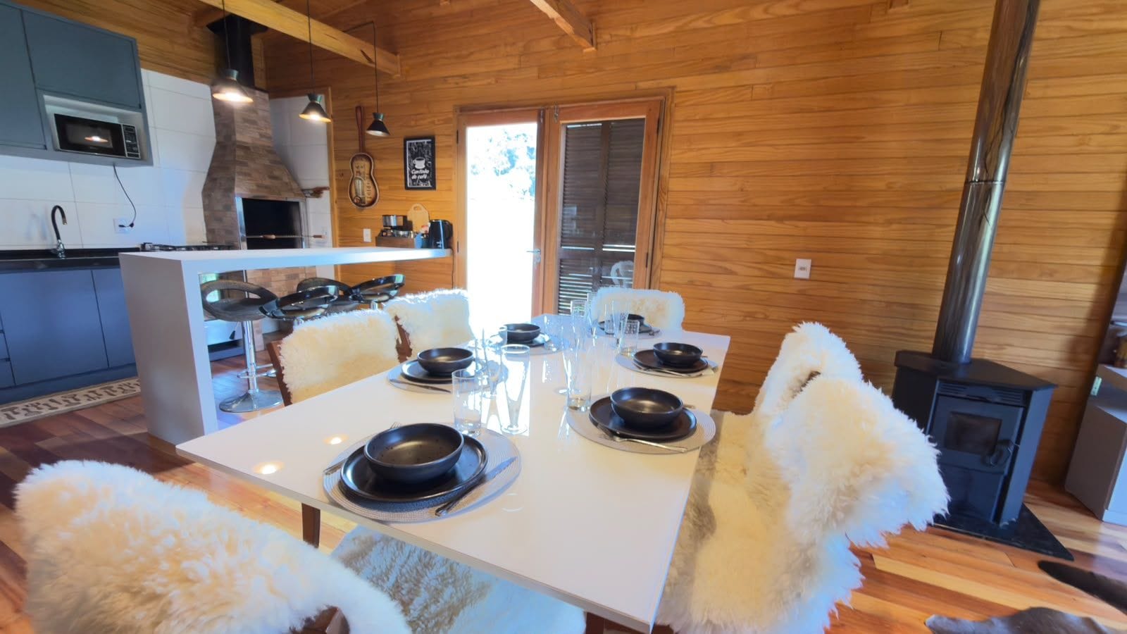 Un comedor con una mesa blanca dispuesta para cuatro personas, rodeada de sillas blancas mullidas. Se observa una estufa de leña al fondo.