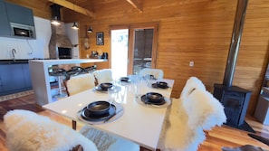 Un comedor con una mesa blanca dispuesta para cuatro personas, rodeada de sillas blancas mullidas. Se observa una estufa de leña al fondo.