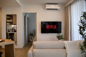 A living room with a flat-screen TV mounted on the wall displaying 'NETFLIX' in red letters, a sofa, a small table, and a potted plant. - Mountain View Corner Unit in Laguna with Rooftop Pool & 10min-walk to the Beach (ตำบลเชิงทะเล)
