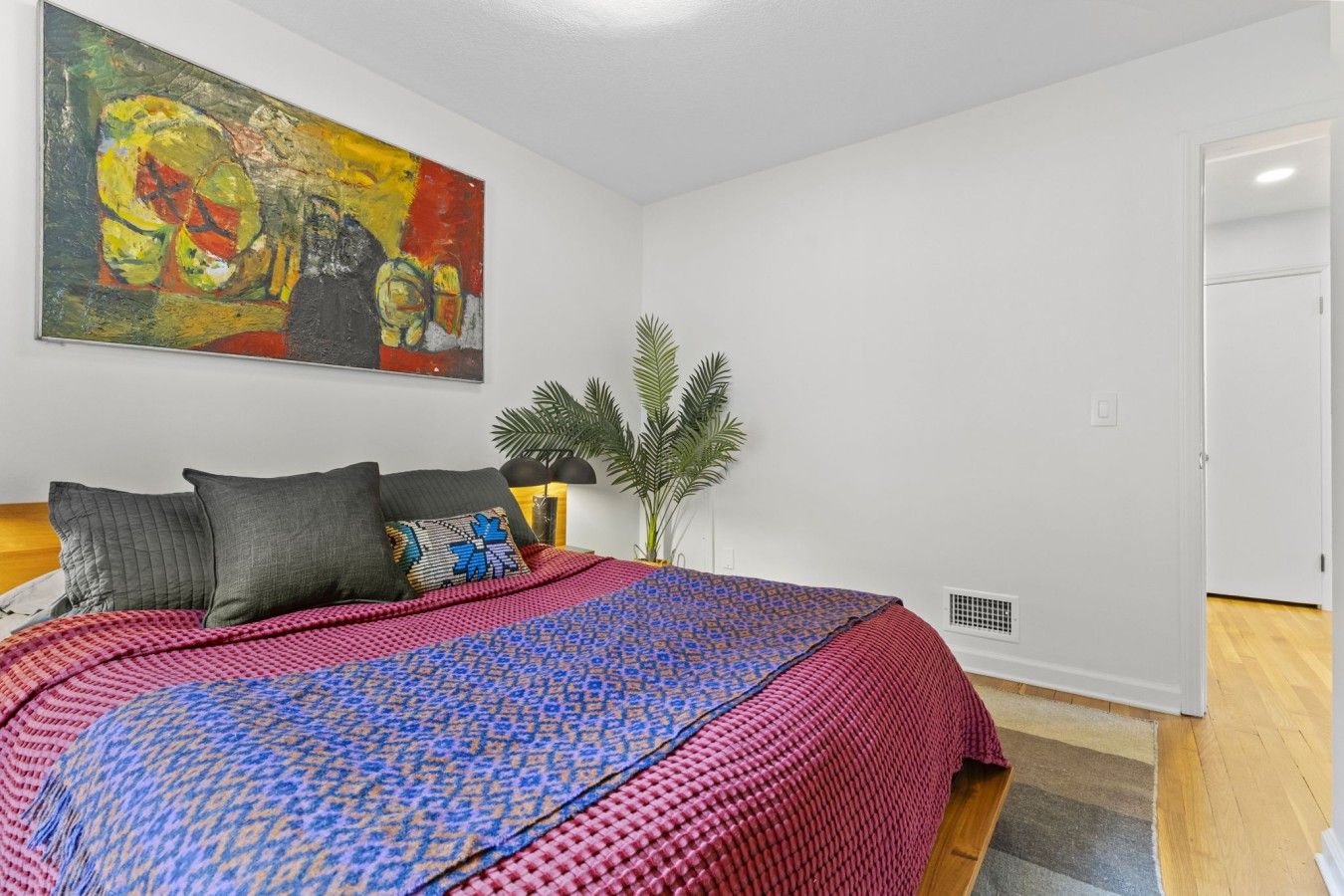 A bedroom with a bed, a painting on the wall, a potted plant, and a door.