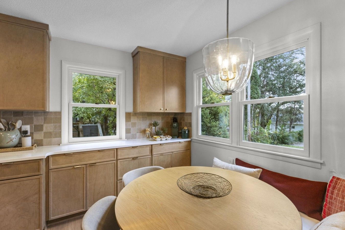 A kitchen with wooden cabinets, a dining table with a woven centerpiece, and a large window with a view of greenery.