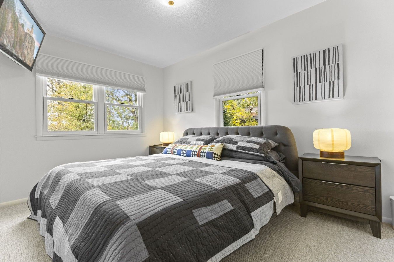Quarto com cama grande, duas mesas de cabeceira, TV fixada na parede e janelas com persianas.