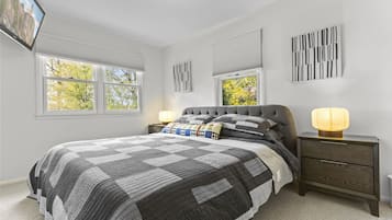 Quarto com cama grande, duas mesas de cabeceira, TV fixada na parede e janelas com persianas.