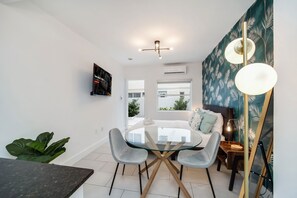 A modern dining area with a glass table, two grey chairs, a dark wooden side table, and a wall-mounted air conditioner. - Byronessa Miami Beach (Miami Beach)