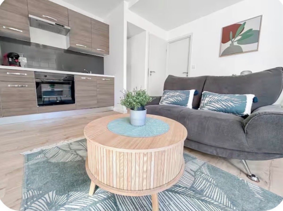 Un espace de vie moderne comprenant une cuisine, un canapé gris et une table basse ronde en bois agrémentée d’une plante.
