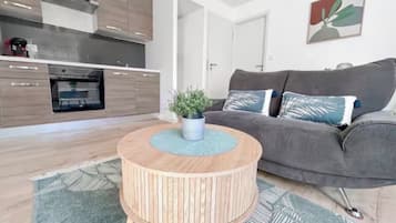 Un espace de vie moderne comprenant une cuisine, un canapé gris et une table basse ronde en bois agrémentée d’une plante.