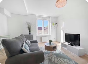 Een moderne woonkamer met een bank, een rond salontafeltje, een televisie en een raam met uitzicht op gebouwen.