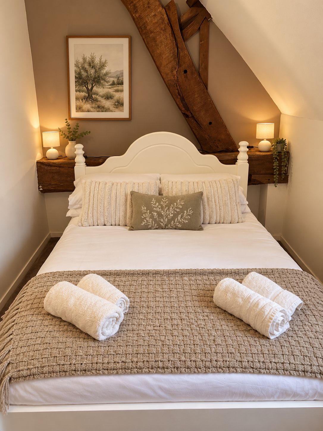 A neatly made bed with a white headboard, two pillows, and a beige blanket. There are two bedside lamps on wooden nightstands, and a framed picture on the wall.