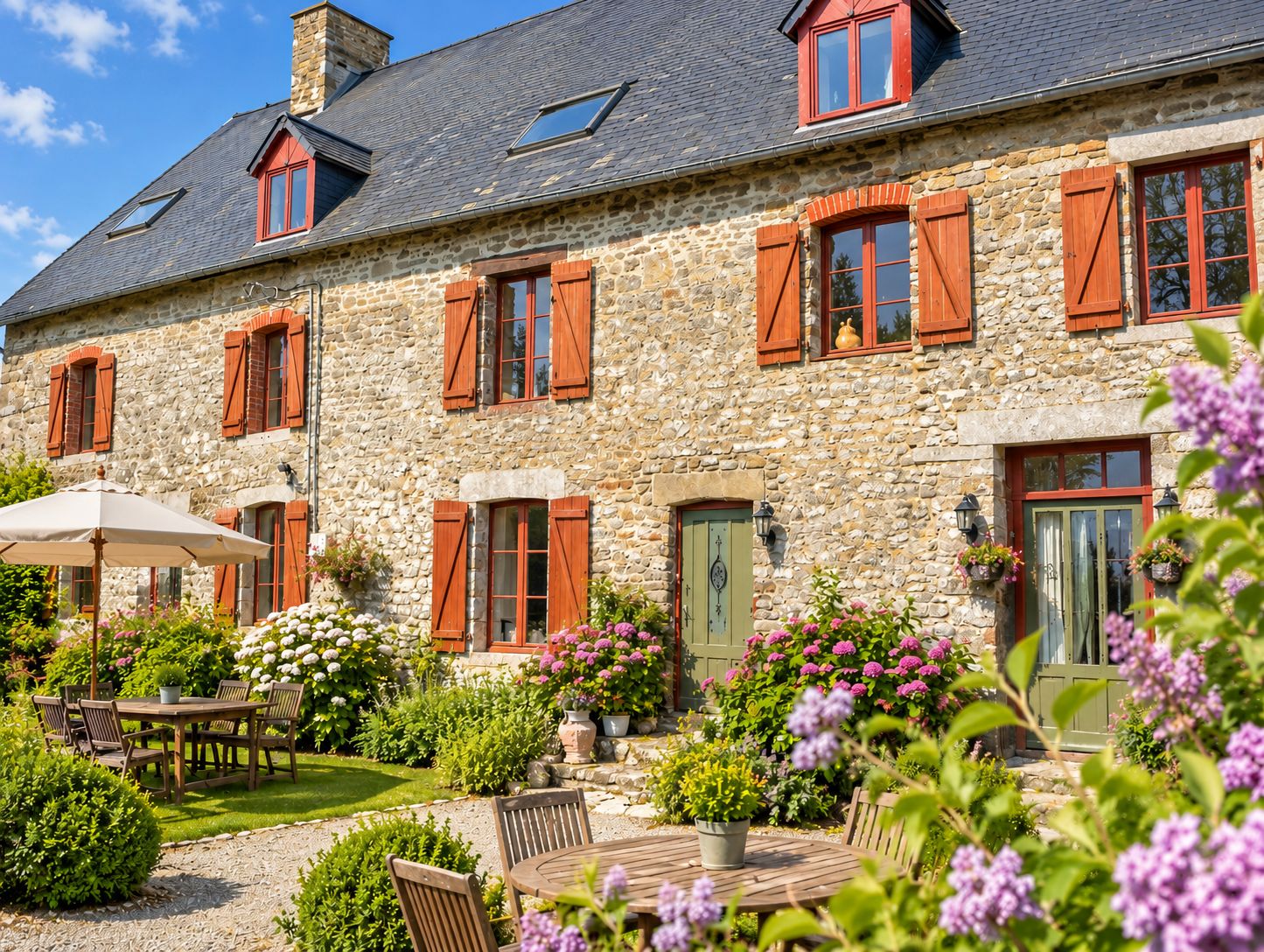 A stone building with multiple windows and shutters, a covered patio with a table and chairs, and a garden with various plants and flowers.