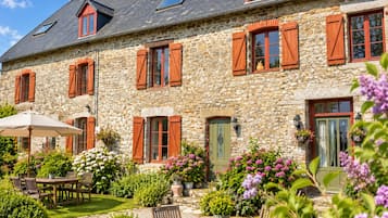 A stone building with multiple windows and shutters, a covered patio with a table and chairs, and a garden with various plants and flowers.