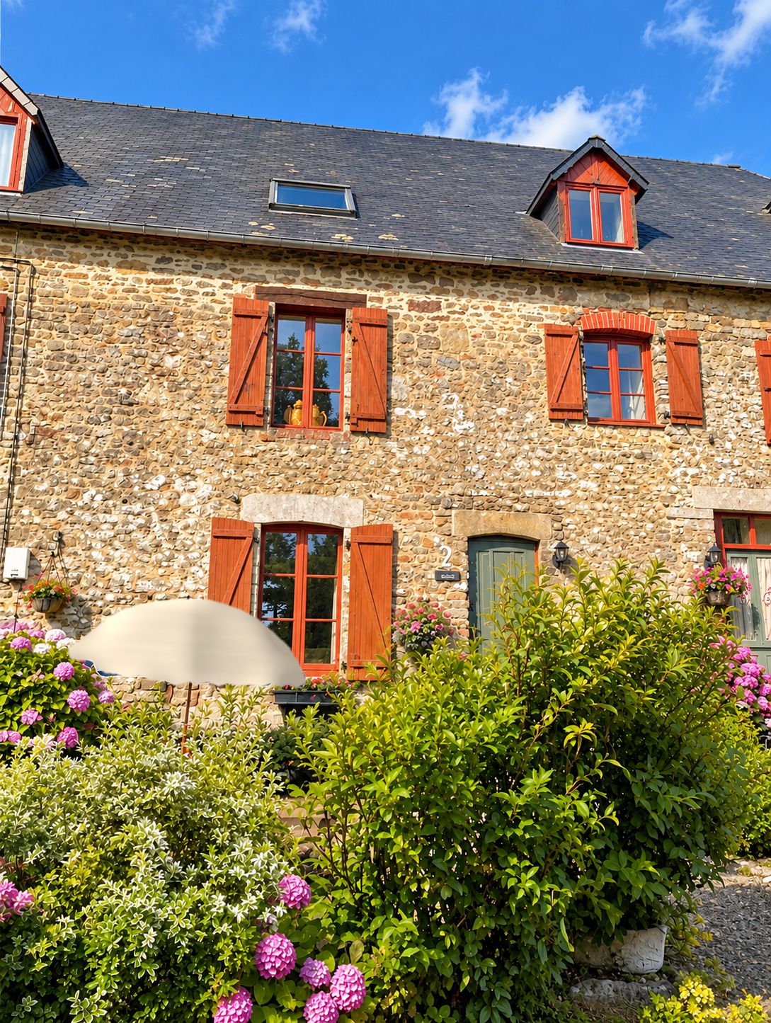 A stone building with multiple windows and shutters, surrounded by lush greenery and flowers.