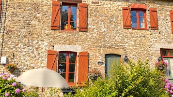 A stone building with multiple windows and shutters, surrounded by lush greenery and flowers.