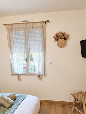 Une chambre à coucher avec un lit, une fenêtre ornée de rideaux légers, un téléviseur fixé au mur et un fauteuil en osier.