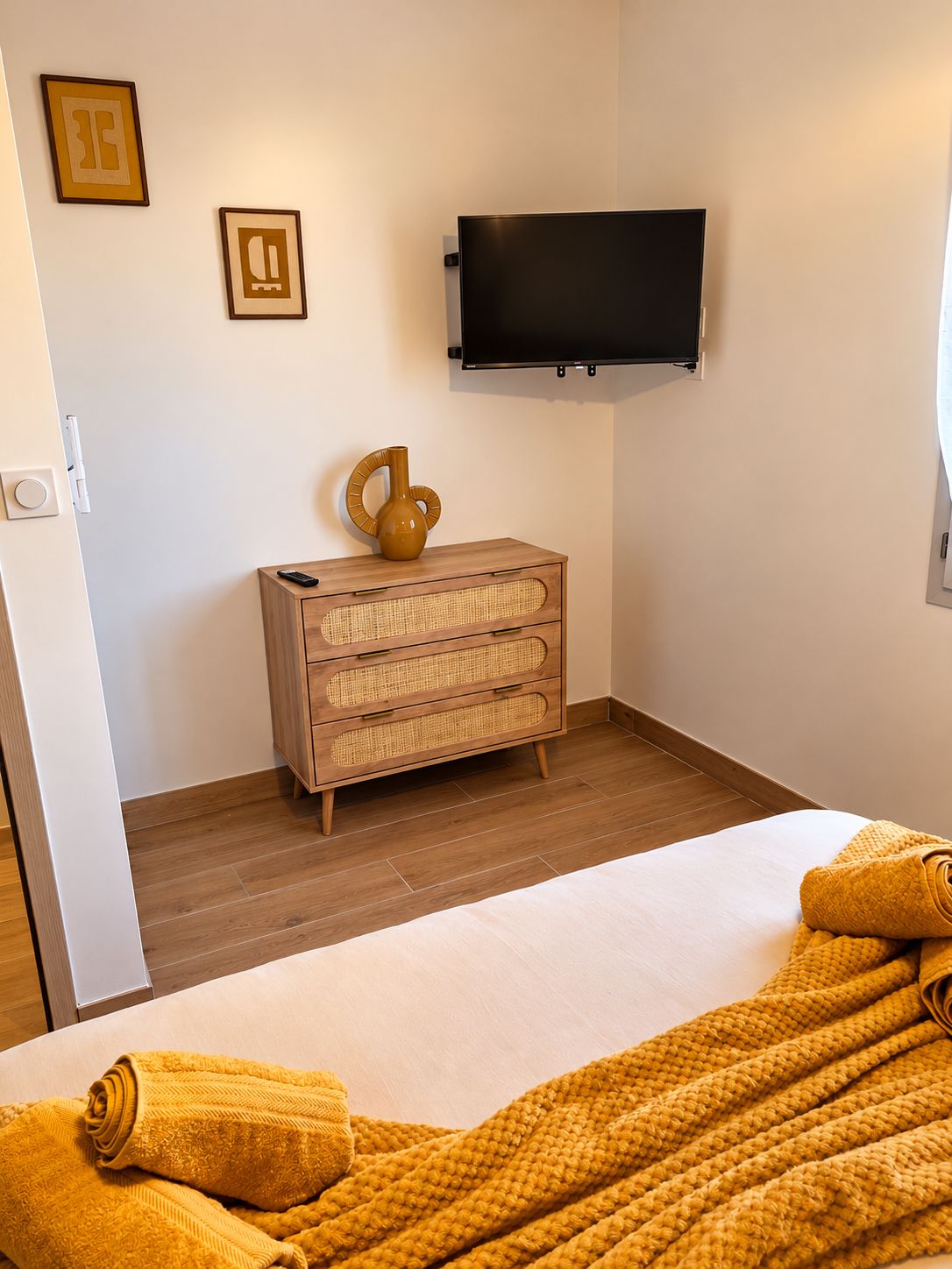 A hotel room with a bed, a wooden dresser, a television mounted on the wall, and two framed pictures on the wall.