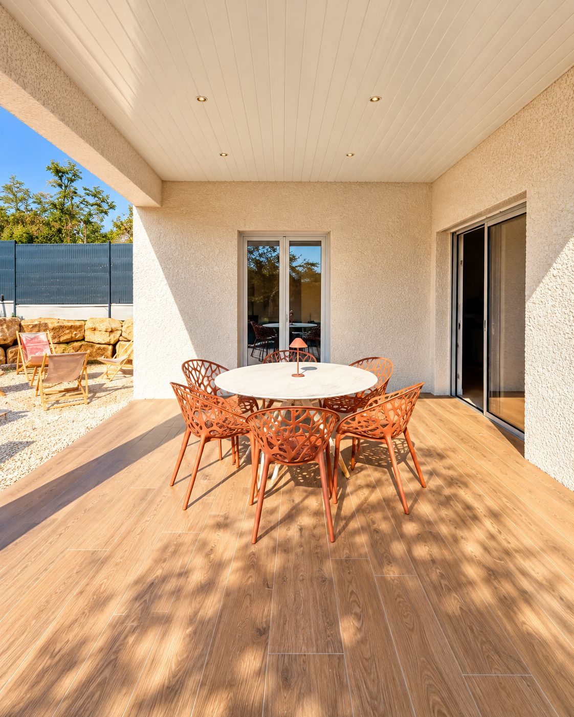 Eine Terrasse im Freien mit einem runden Tisch und orangefarbenen Stühlen, einem Holzboden und Blick ins Grüne.