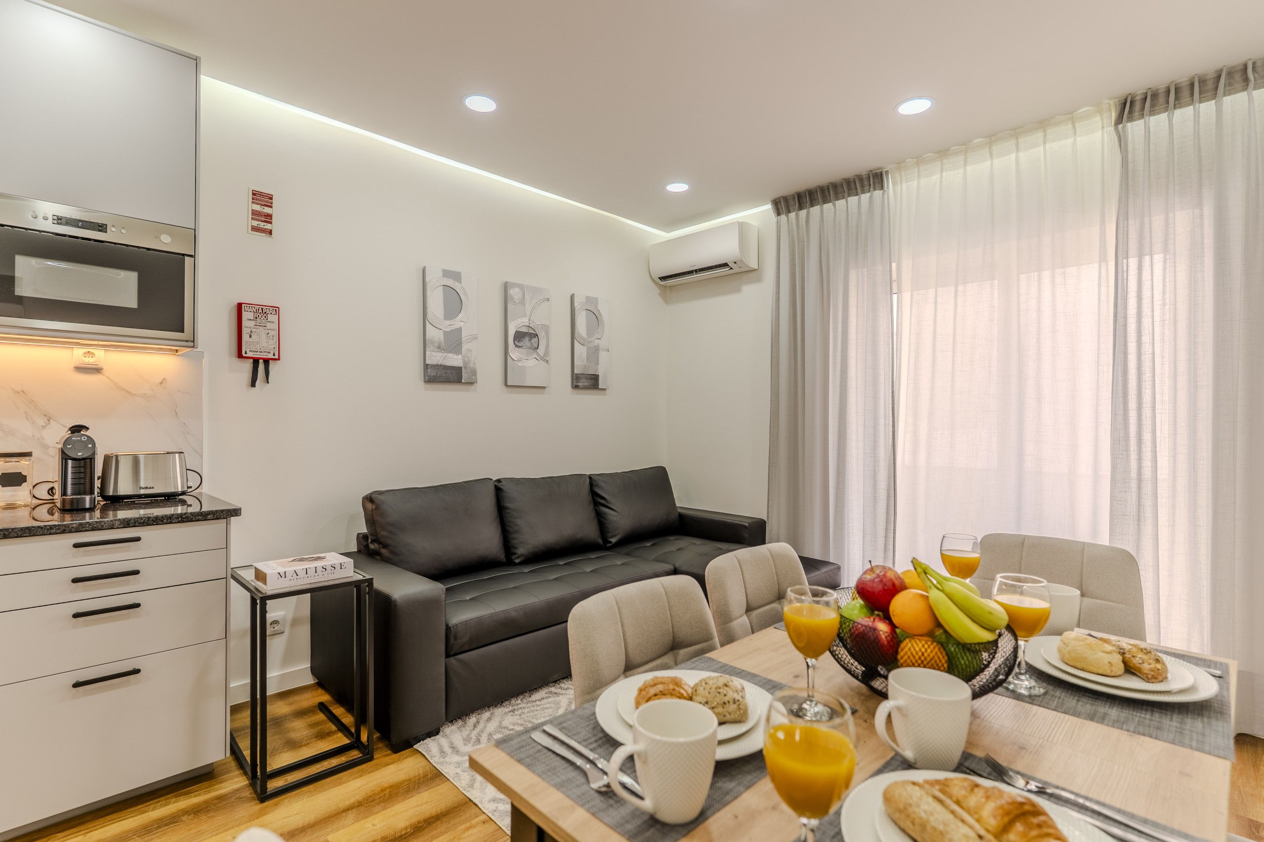 A modern kitchen with a microwave, toaster, and coffee maker. There is a dining area with a table set for two, featuring plates, glasses, and a fruit bowl. A black leather sofa is against the wall, and there are three framed artworks above it.