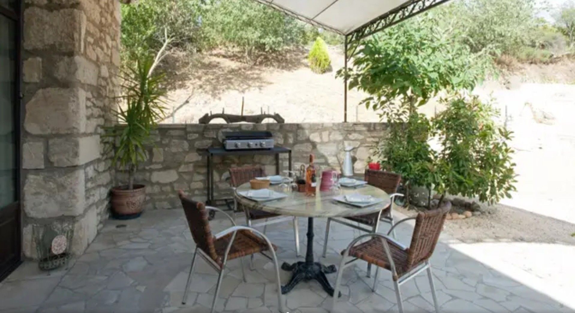 Eine Terrassenausstattung mit Steinwand, einem runden Tisch, vier Korbstühlen, einem Grill und Topfpflanzen.
