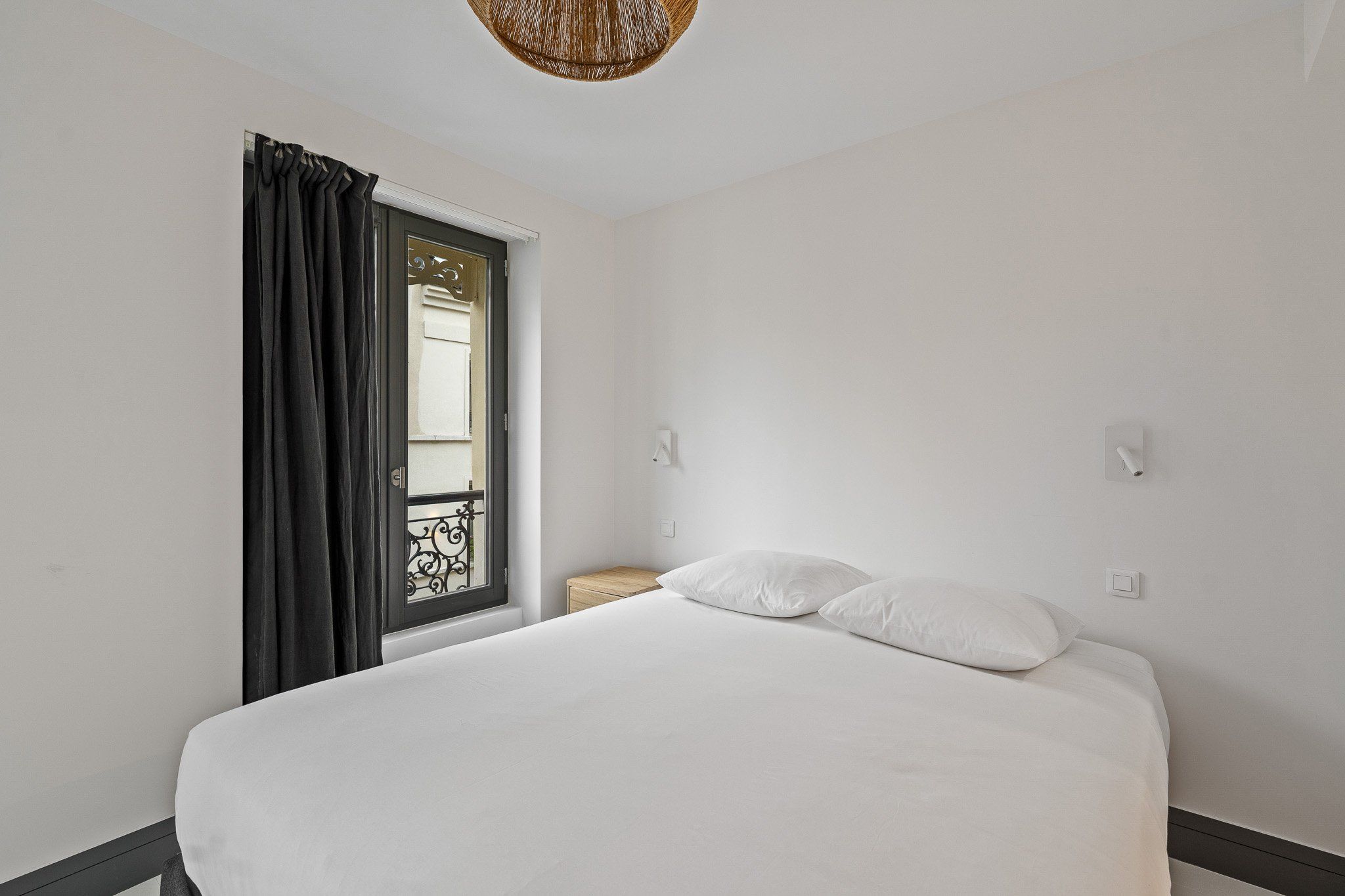 A bedroom with a bed, white bedding, a window with curtains, a nightstand, and a wall-mounted light switch.