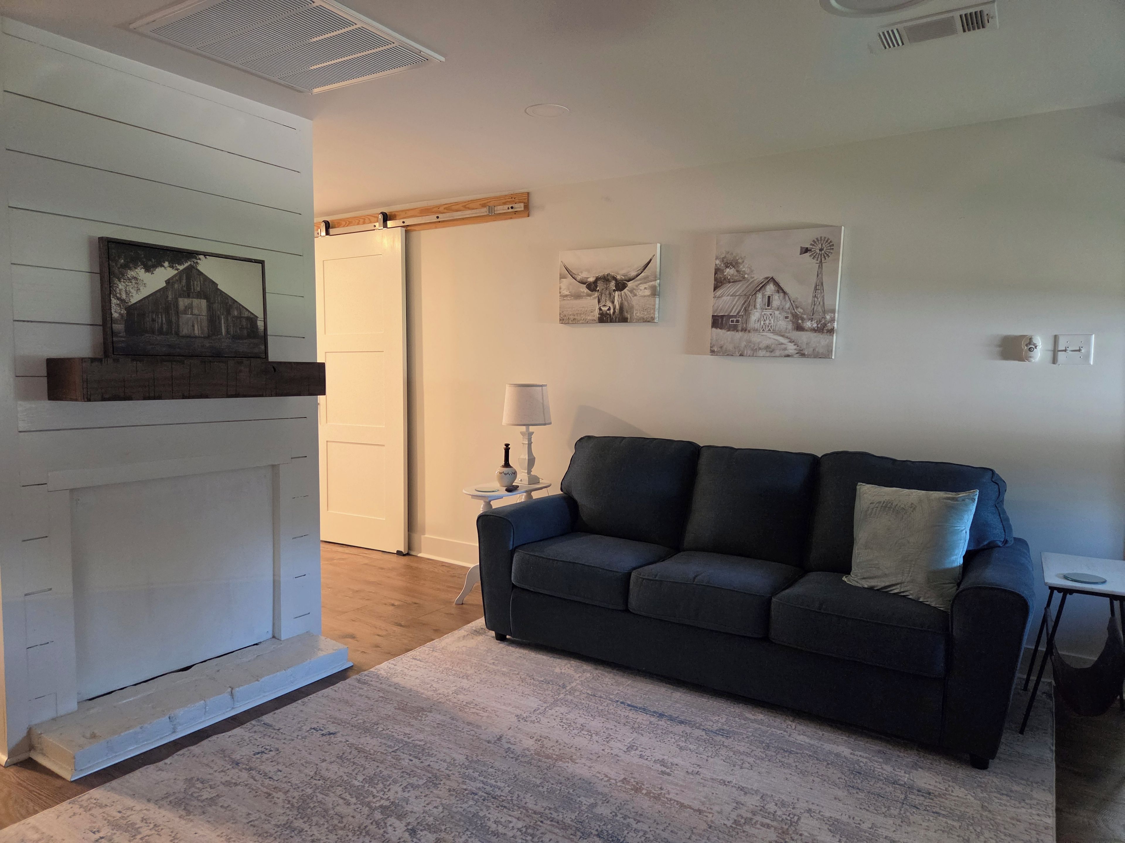 A room with a dark blue sofa, a small round table with a lamp, and two wall-mounted pictures depicting farm scenes.