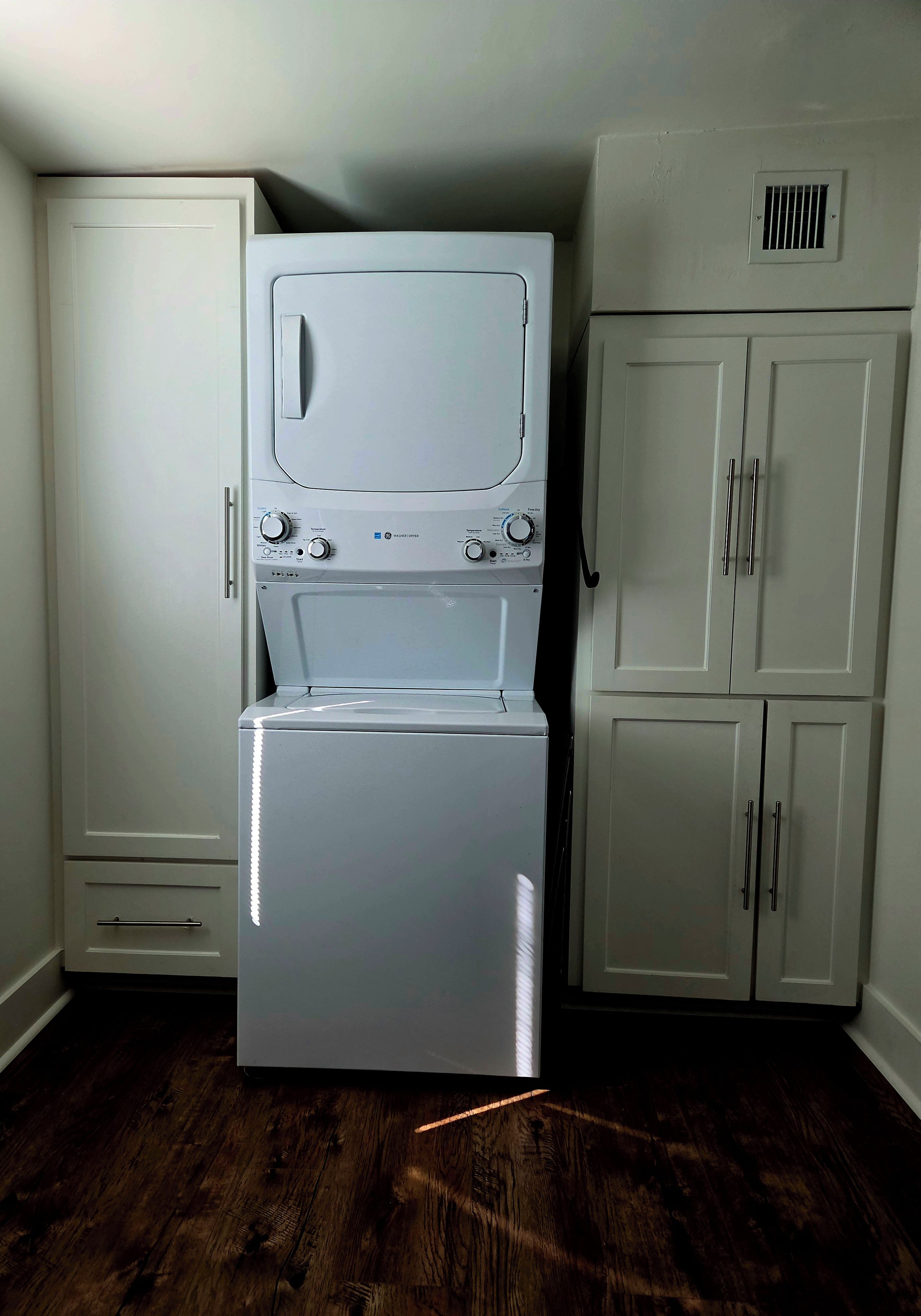 A laundry room with a front-loading washer and dryer, a white cabinet with a vent, and wooden flooring.