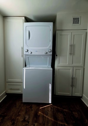 A laundry room with a front-loading washer and dryer, a white cabinet with a vent, and wooden flooring.