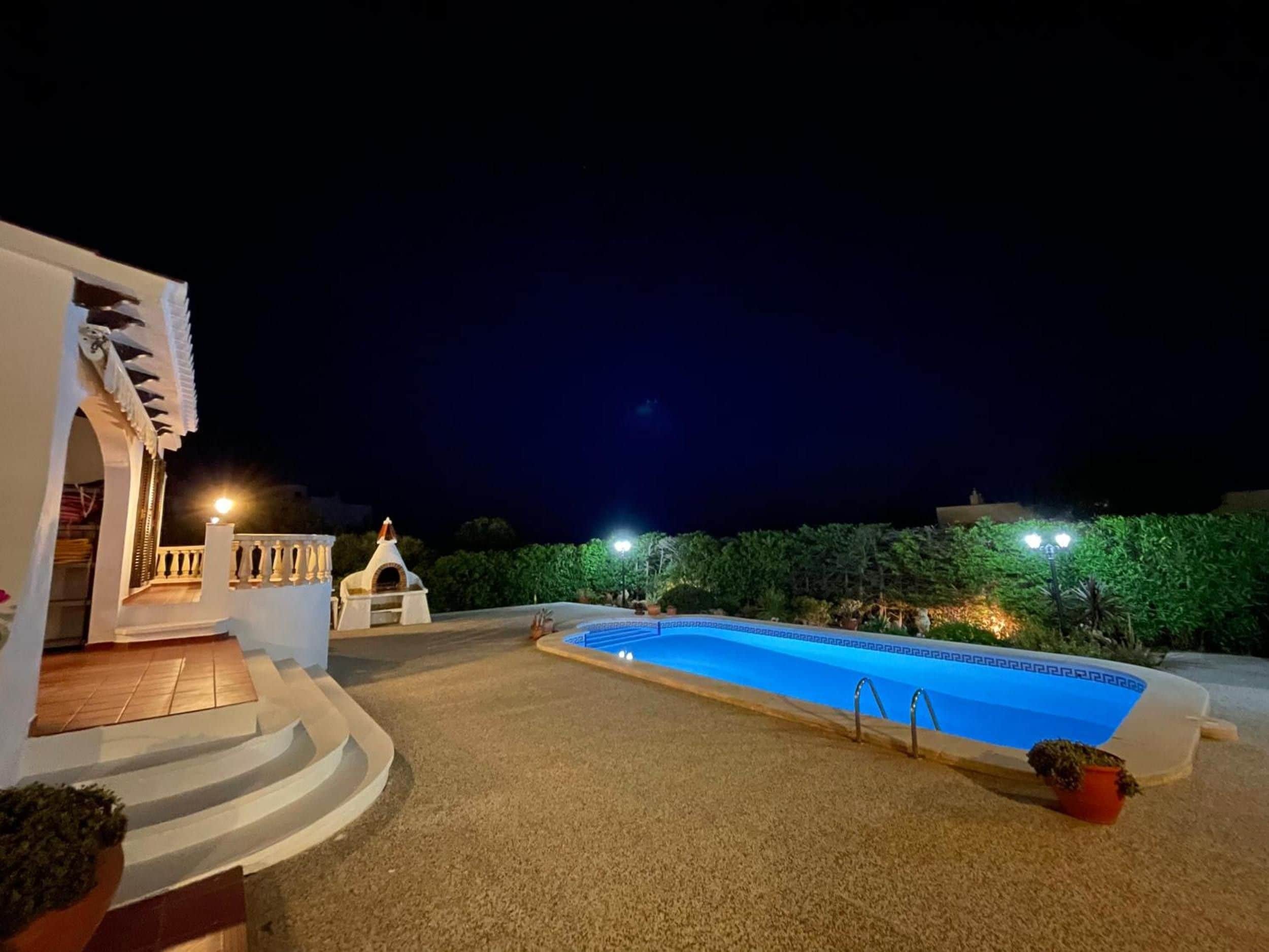 A swimming pool with a white deck, a small building with a conical roof, and a well-lit night sky.