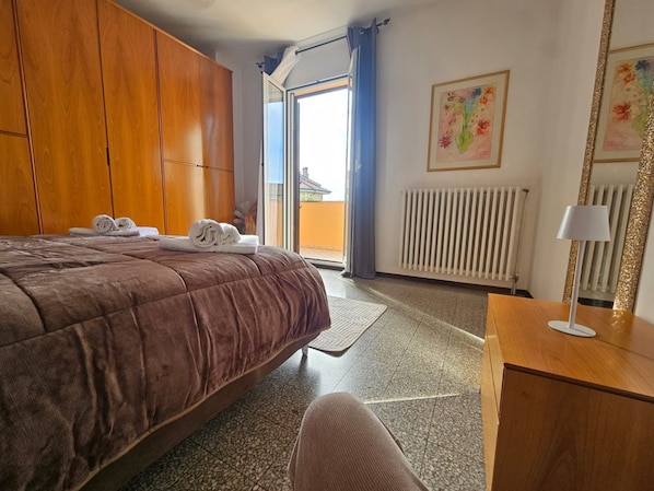 Une chambre d’hôtel avec un grand lit, une armoire en bois, un radiateur, un bureau avec une lampe et une fenêtre avec des rideaux.
